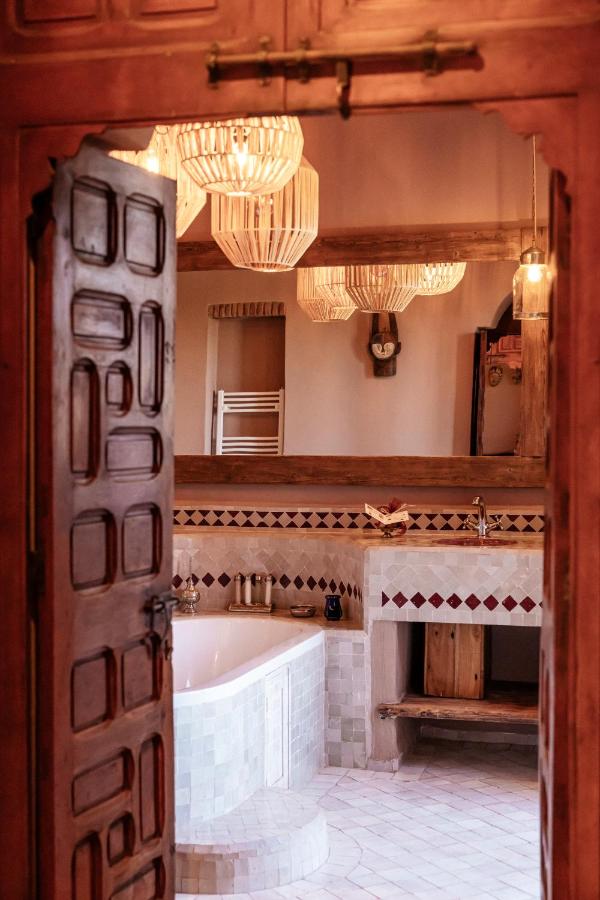 10 BEST Riads in Essaouira 51 A rustic bathroom reminiscent of riads in Essaouira, with a wooden door partially open, revealing a curved bathtub, tiled walls with diamond patterns, wooden beams, a bench, and several hanging lantern-style lights.