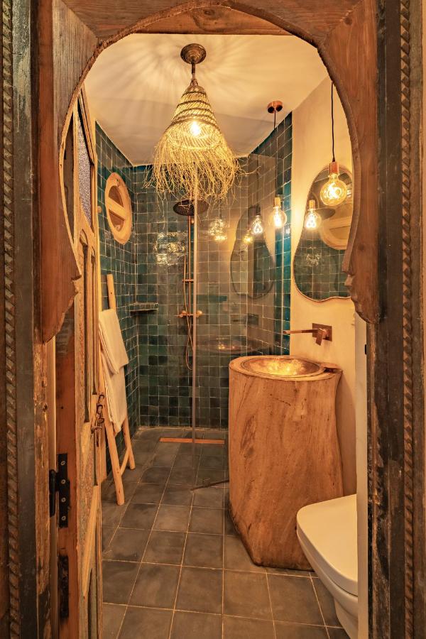 10 BEST Riads in Essaouira 42 A cozy bathroom with rustic wooden accents and green tiled walls reminiscent of riads in Essaouira features a round wooden sink, walk-in shower, hanging pendant lights, a decorative arched doorway, and a towel ladder against the wall.