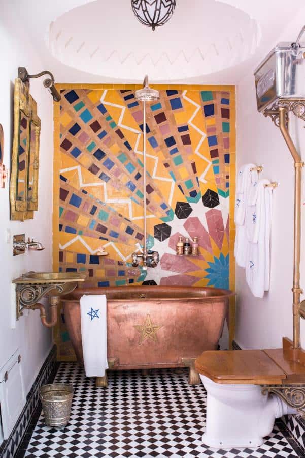 10 BEST Riads in Essaouira 26 A colorful bathroom reminiscent of riads in Essaouira, featuring a copper bathtub, mosaic tile wall in geometric patterns, black-and-white checkered floor, vintage fixtures, and white towels with blue star designs.