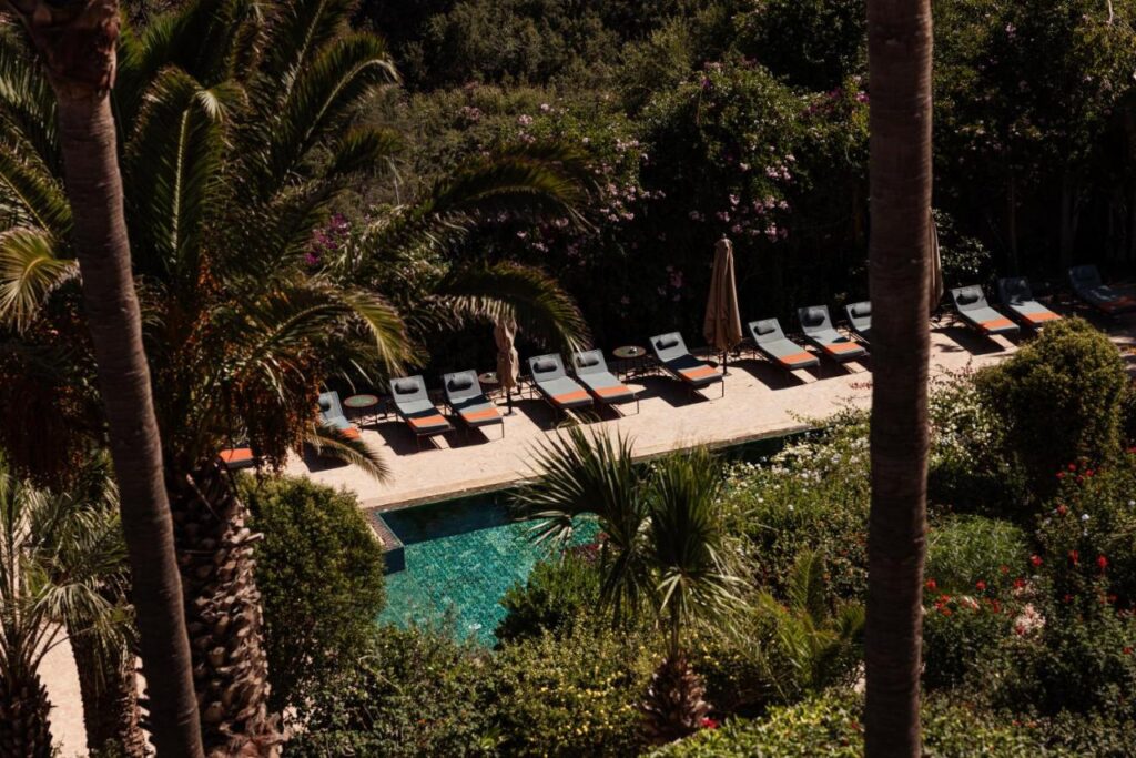 10 BEST Riads in Essaouira 48 A row of empty lounge chairs and umbrellas lines the edge of a turquoise pool, surrounded by lush greenery and palm trees, creating a serene, tropical atmosphere reminiscent of riads in Essaouira.