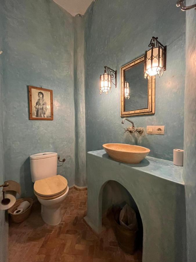 10 BEST Riads in Essaouira 38 A small bathroom with textured blue walls, a white toilet with a wooden seat, a stone sink on a blue countertop, a mirror, lantern-style wall lights, and a framed photo evokes the charm of riads in Essaouira.