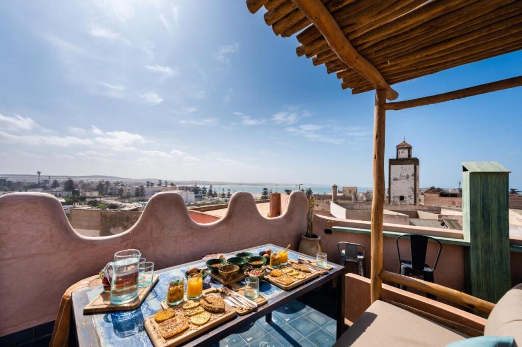 10 BEST Riads in Essaouira 41 A rooftop terrace, reminiscent of riads in Essaouira, features a wooden pergola overlooking the cityscape and sea. A table set for breakfast with plates, bread, juice, and coffee invites you under a blue sky dotted with clouds.