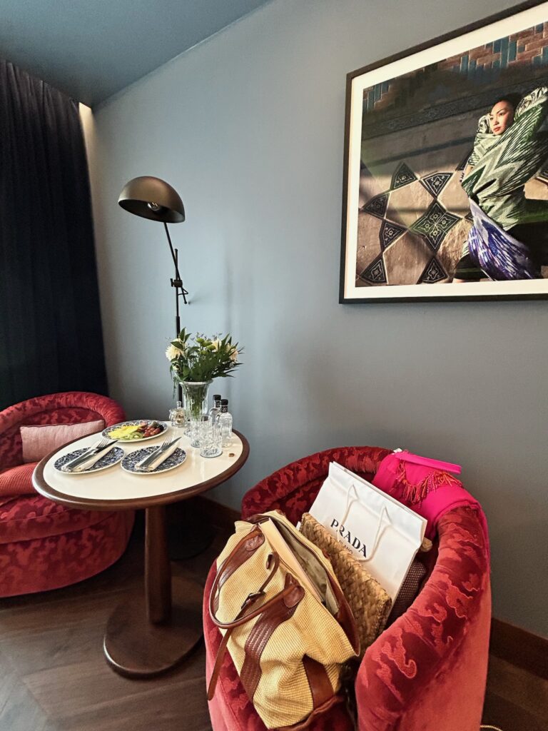 A cozy seating area with two red velvet chairs, a round table set with dishes and glassware—perfect for planning your Southern Spain itinerary 10 days—plus a floor lamp, framed photo, and shopping bags and clothing draped over one chair.