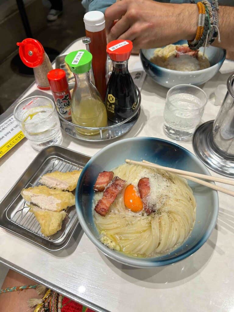 ULTIMATE 10 Day Japan Travel Itinerary 6 A bowl of noodles topped with an egg yolk, grated cheese, and bacon sits on a table next to fried food, sauces, and water—an inviting scene you might find during your 10 day Japan travel itinerary. A hand with bracelets holds chopsticks.