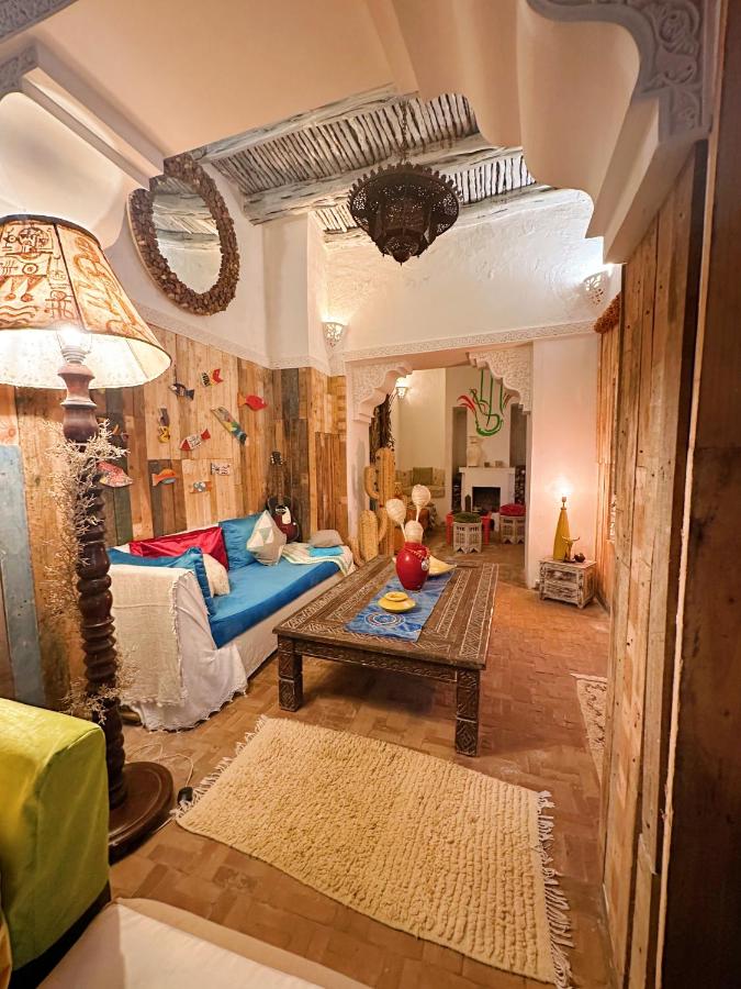 10 BEST Riads in Essaouira 47 Cozy living room with rustic wooden walls, a carved wooden coffee table, sofas with colorful cushions, a large ornate lamp, a decorative mirror, and Moroccan-style decor reminiscent of riads in Essaouira under a white arched ceiling.