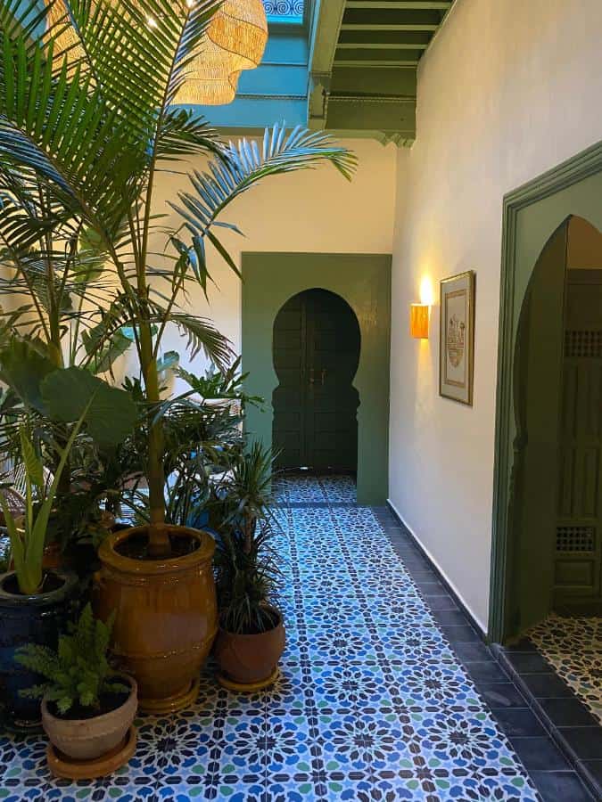 10 BEST Riads in Essaouira 12 Indoor hallway inspired by riads in Essaouira, with colorful patterned tiles, green arched doorways, potted plants in terracotta pots, and a wall-mounted light. Ceiling features blue beams and a yellow decorative shade overhead.