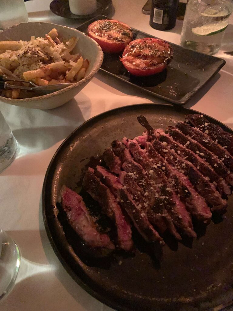 15 Most Fun Restaurants in Madrid 47 A plate of sliced steak topped with grated cheese, a bowl of seasoned fries, and two baked tomato halves on a dark table at one of the fun restaurants in Madrid.