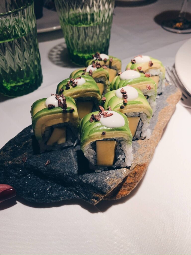 15 Most Fun Restaurants in Madrid 42 A platter of sushi rolls topped with avocado slices, white sauce, and small brown sprinkles, arranged neatly on a stone serving board—just one of the delights you’ll find at fun restaurants in Madrid, with two green textured glasses in the background.