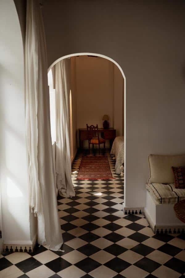 10 BEST Riads in Essaouira 16 A hallway reminiscent of riads in Essaouira features black-and-white checkered tile flooring, white walls, and an arched doorway leading to a cozy room with a wooden desk, chair, and red patterned rug. Sunlight filters in, casting soft shadows.