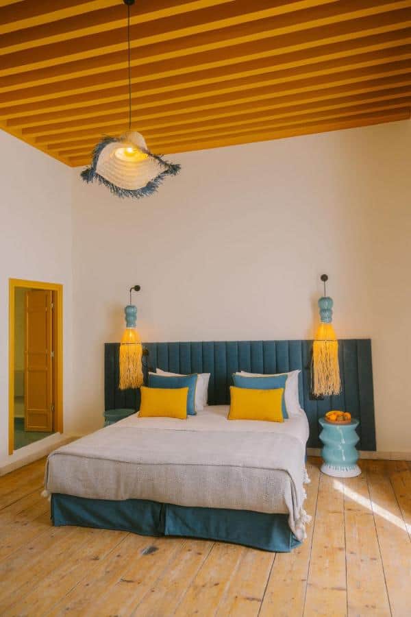 10 BEST Riads in Essaouira 11 A cozy bedroom inspired by riads in Essaouira, with a blue bed, yellow and gray pillows, hanging fringe lamps, a blue nightstand with oranges, wooden floors, white walls, and a ceiling with exposed wooden beams.
