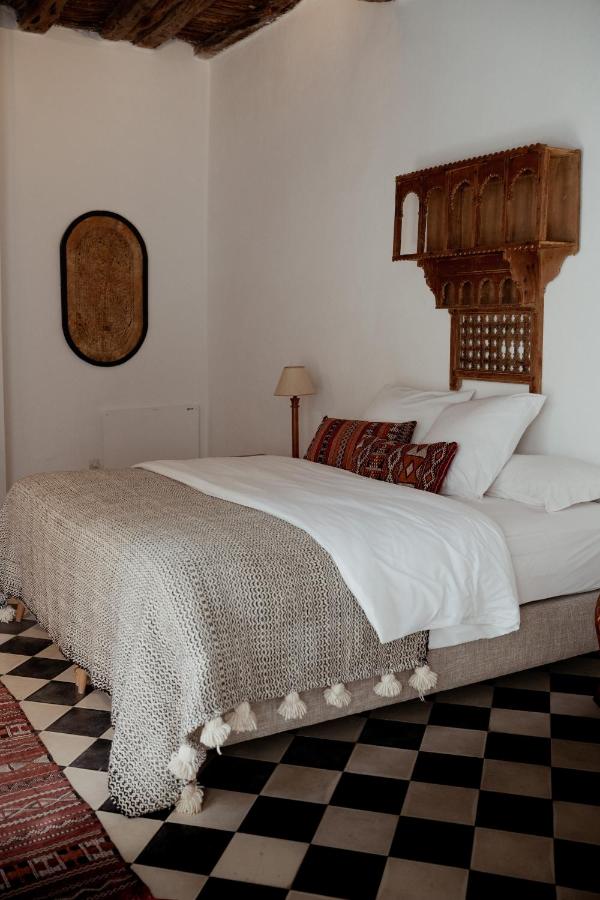 10 BEST Riads in Essaouira 19 A cozy bedroom inspired by riads in Essaouira, with a bed covered in white linens, textured blanket, and patterned pillows. The room features checkerboard floor tiles, a wooden wall shelf, standing lamp, and rustic decor on white walls.