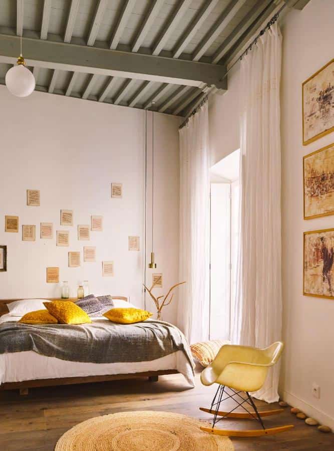10 BEST Riads in Essaouira 20 A cozy bedroom inspired by riads in Essaouira, featuring a wooden bed, yellow cushions, wall art, high gray beamed ceiling, white curtains, a beige rug, and a cream Eames-style chair by a sunlit window.