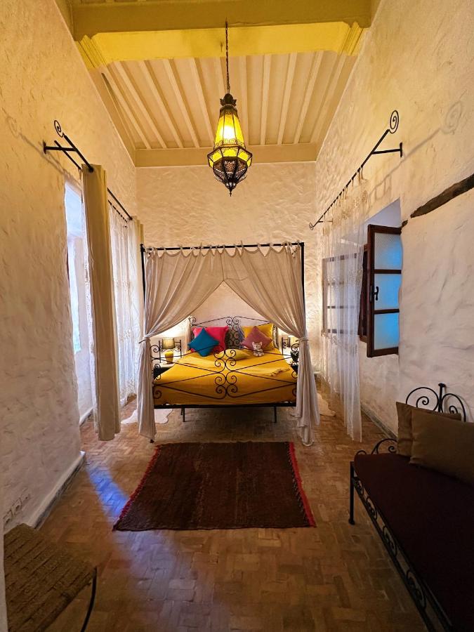 10 BEST Riads in Essaouira 45 A cozy bedroom reminiscent of riads in Essaouira, featuring white textured walls, a yellow canopied bed with colorful pillows, a hanging lantern, open windows, and a red rug on a wooden floor.