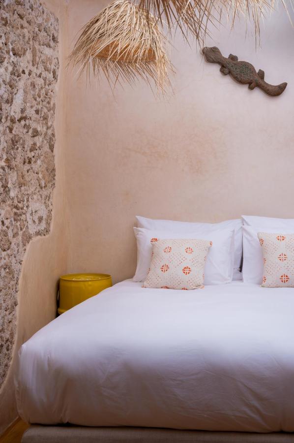 10 BEST Riads in Essaouira 40 A cozy bedroom inspired by riads in Essaouira, featuring a white bed, patterned pillows, a yellow side table, a textured stone wall, and a decorative lizard on the pale wall. Dried grass decor hangs from the ceiling.