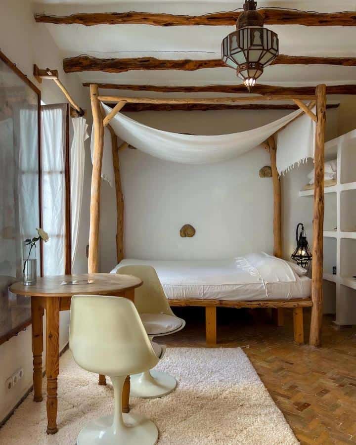 10 BEST Riads in Essaouira 37 A cozy bedroom inspired by riads in Essaouira, featuring a rustic wooden canopy bed, white bedding, a round wooden table, two modern white chairs, a textured rug, exposed beams, and a hanging lantern-style light fixture.