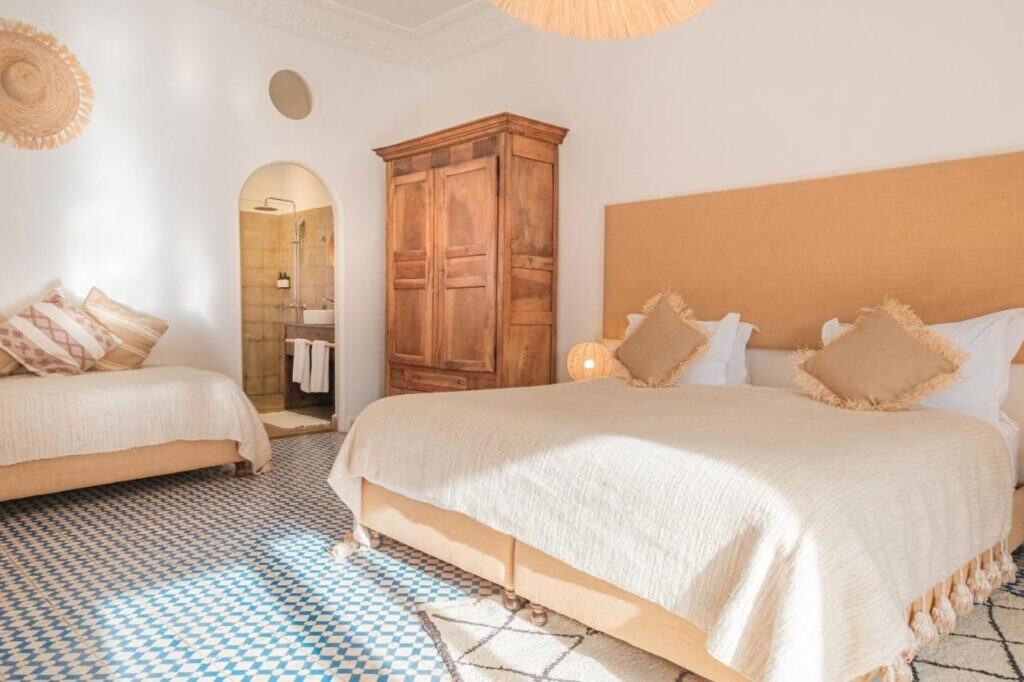 10 BEST Riads in Essaouira 30 Bright bedroom in the style of riads in Essaouira, featuring a large bed and a single bed with beige bedding. A wooden wardrobe, patterned tile floor, and open doorway to the bathroom complete this naturally lit space.