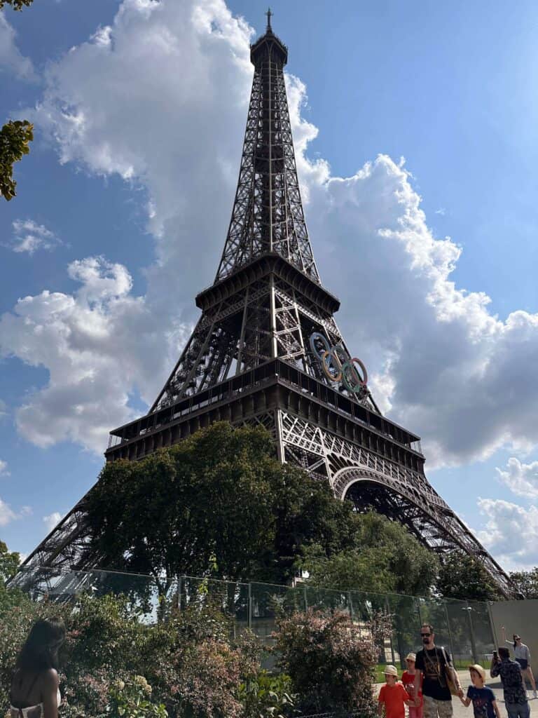 The Eiffel Tower stands tall against a partly cloudy sky, Olympic rings on display—a must-see for those seeking things to do in Paris with teenagers. People walk in the foreground among trees and greenery.