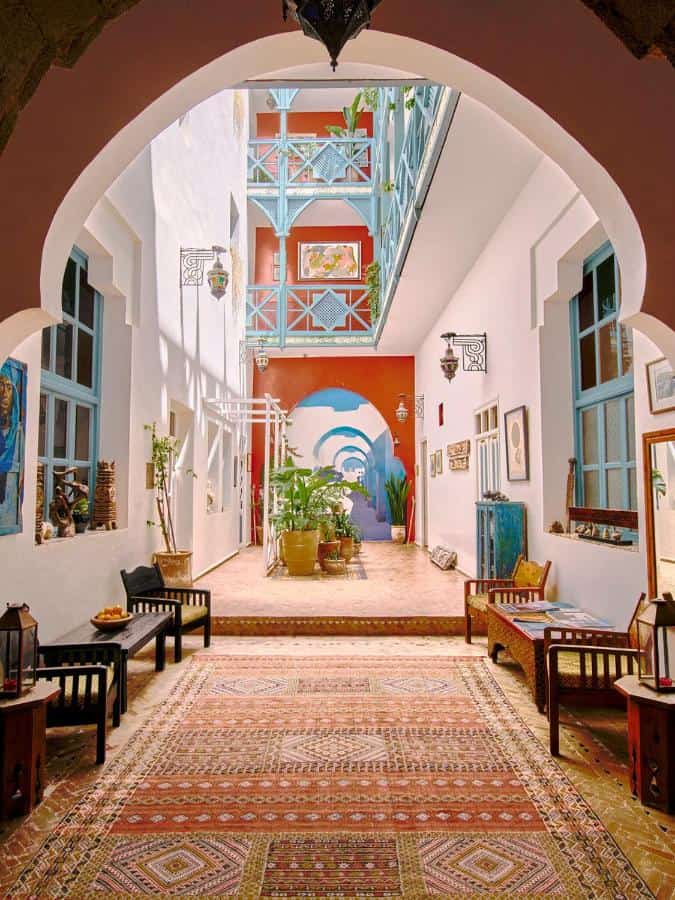 10 BEST Riads in Essaouira 34 A bright, colorful interior courtyard inspired by riads in Essaouira, with patterned rugs, wooden benches, potted plants, and blue balconies. Arched doorways and lattice windows create a vibrant, light-filled space.