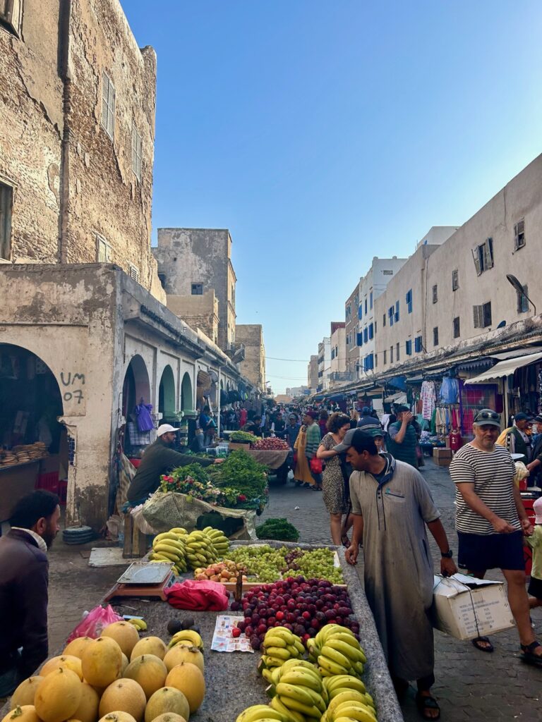 10 BEST Riads in Essaouira 6 A bustling outdoor market lined with old buildings and riads in Essaouira, where vendors sell fresh fruits and vegetables while shoppers browse stalls under a clear blue sky.