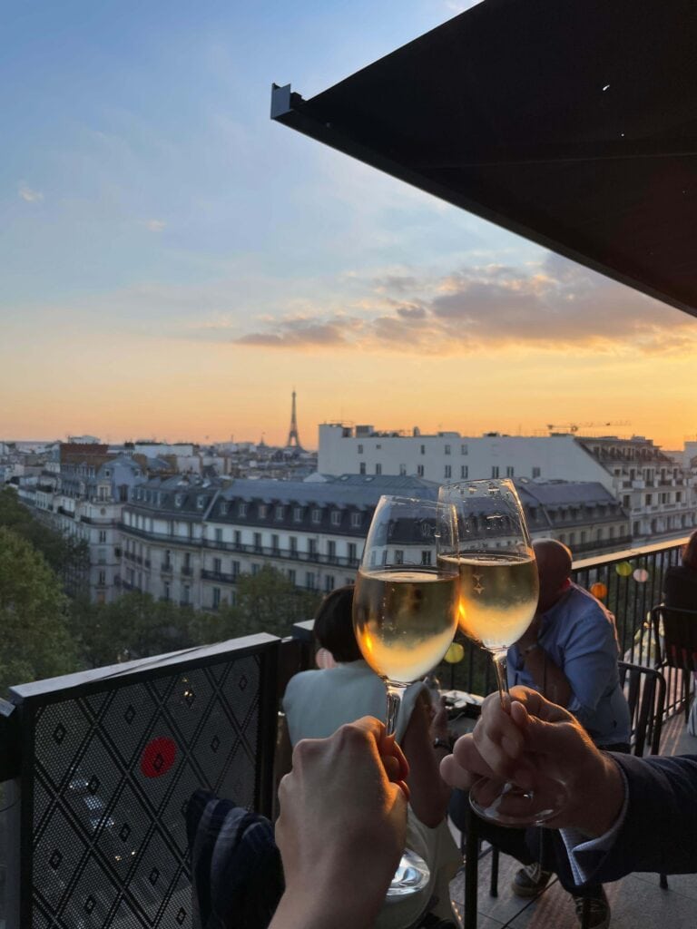 13 Unique Hotels in Paris for a Stylish Stay 3 Two people clinking glasses of white wine on a balcony at sunset, overlooking Paris rooftops with the Eiffel Tower in the background—a perfect end to a day exploring things to do in Paris with teenagers.