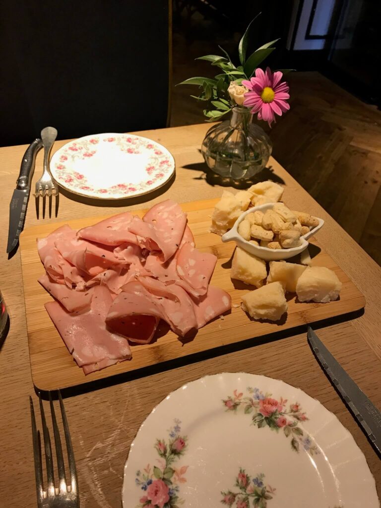 15 Most Fun Restaurants in Madrid 44 A wooden board with sliced deli meat, chunks of cheese, and a small dish of croutons sits on a table set with floral plates and cutlery—perfect for those seeking fun restaurants in Madrid with charming décor.