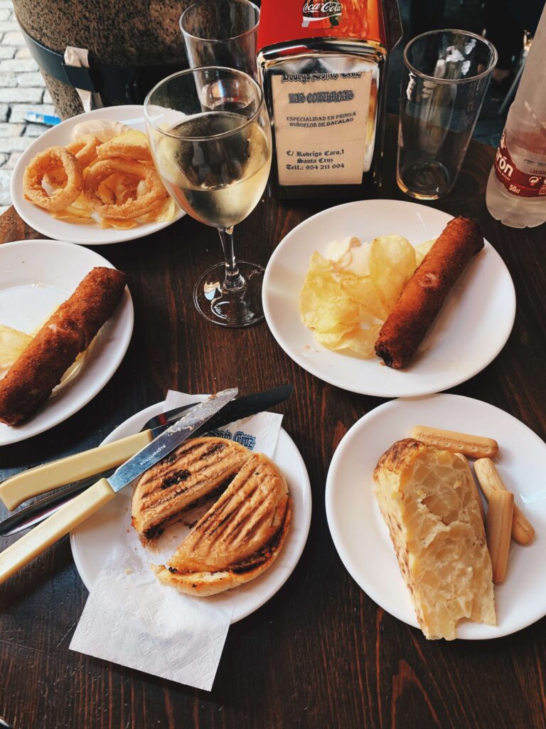 Southern Spain Itinerary 10 Days 12 A table set with assorted Spanish tapas—croquettes, onion rings, potato chips, bread, breadsticks, a toasted sandwich—plus a glass of white wine and bottled water on small plates; perfect inspiration for your Southern Spain itinerary 10 days.