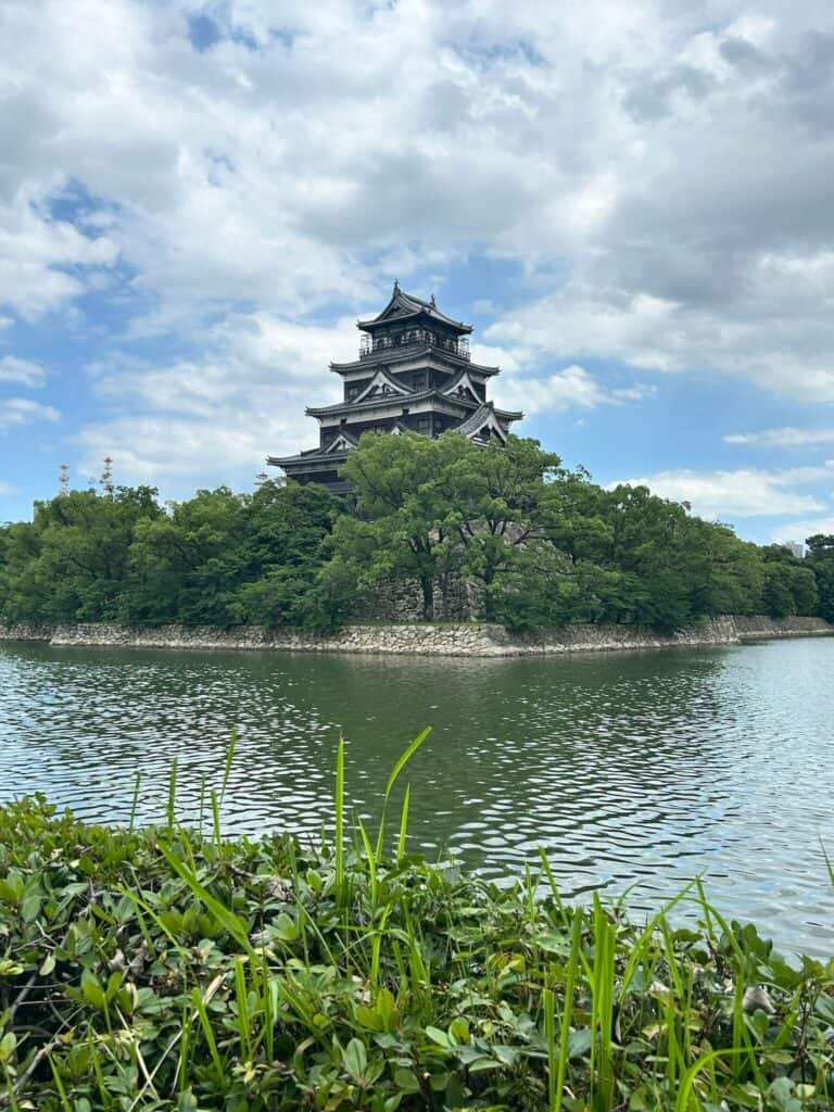ULTIMATE 10 Day Japan Travel Itinerary 18 A traditional Japanese castle stands surrounded by trees on an island, its stone wall at the base reflected in calm water—a must-see highlight on any 10 day Japan travel itinerary under a partly cloudy sky.