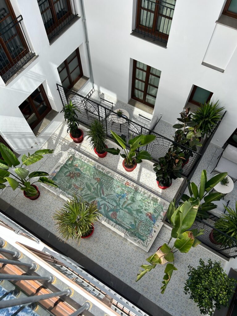 A courtyard viewed from above, featuring potted green plants around a rectangular, mosaic-tiled area with floral and fish designs—perfect inspiration for your Southern Spain itinerary 10 days. The white building has dark window frames and balconies.