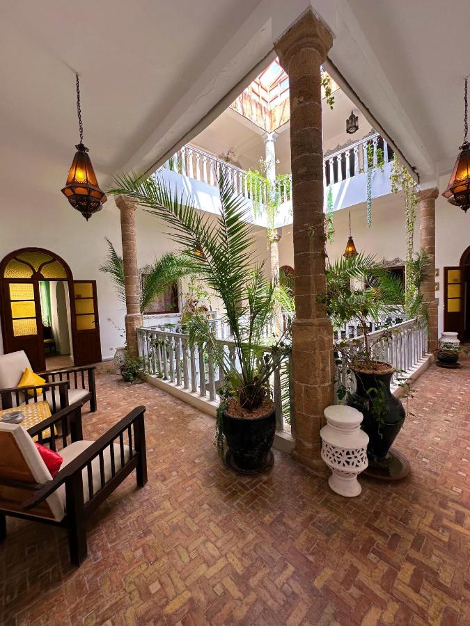 10 BEST Riads in Essaouira 44 A bright indoor courtyard, reminiscent of riads in Essaouira, with potted plants, stone columns, wooden chairs with cushions, hanging lanterns, and a balcony above, featuring natural light from an open ceiling.