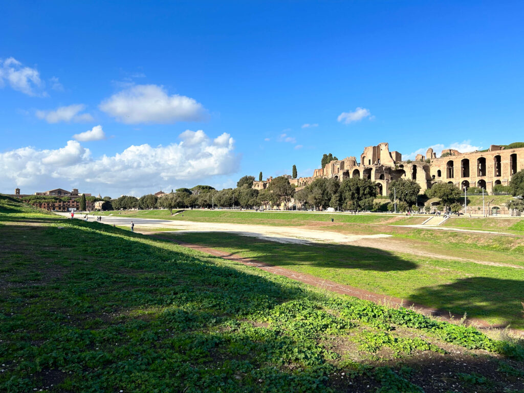 3 Days in Rome Itinerary 7 The image shows the ancient Circus Maximus in Rome, Italy—an iconic site often featured in a 3 days in Rome itinerary—with grassy areas, a partially dry track, ruins, and trees under a bright blue sky with scattered clouds.