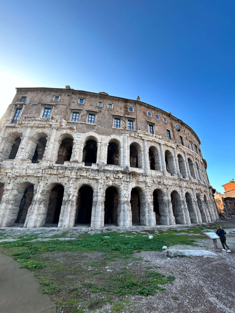 3 Days in Rome Itinerary 6 Ancient stone amphitheater with multiple arches and columns, viewed from below against a clear blue sky—a must-see stop on any 3 days in Rome itinerary. A person stands near an information board, with green grass and dirt in the foreground.