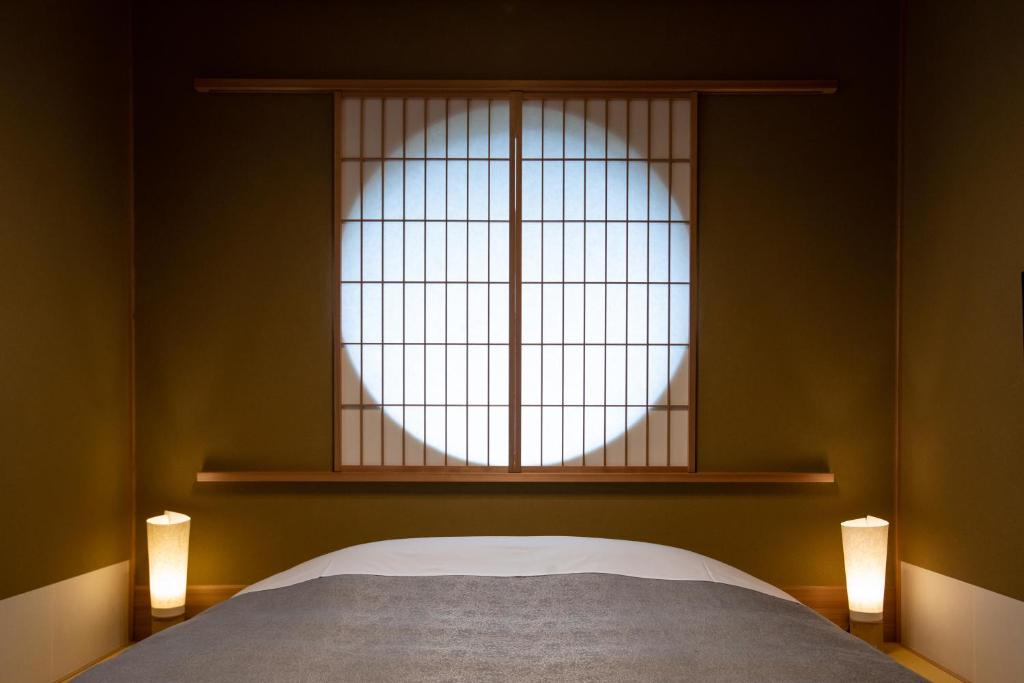 8 UNIQUE Miyajima Ryokans, Hiroshima 37 A minimalist Japanese-style bedroom reminiscent of Miyajima ryokans, with a futon, shoji screen window casting a circular light pattern, and two soft-glowing lamps on either side of the bed.