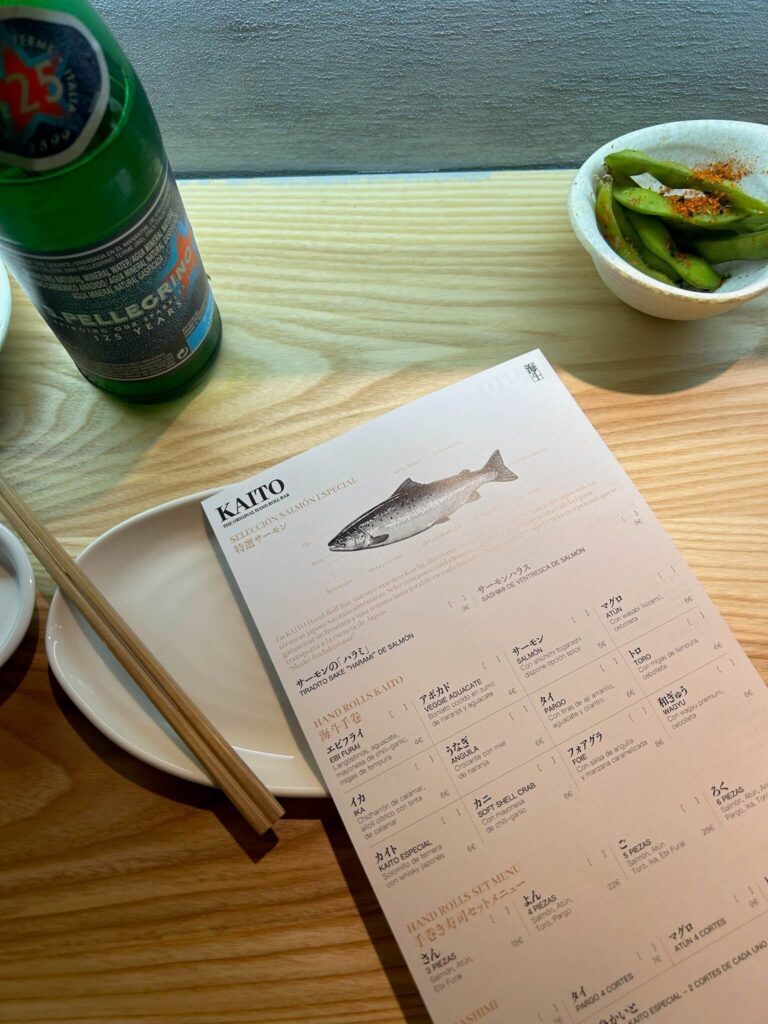 15 Most Fun Restaurants in Madrid 40 A restaurant table with a menu on a plate, wooden chopsticks, a bottle of San Pellegrino sparkling water, and a bowl of seasoned edamame on a light wooden surface—perfect for those seeking fun restaurants in Madrid.