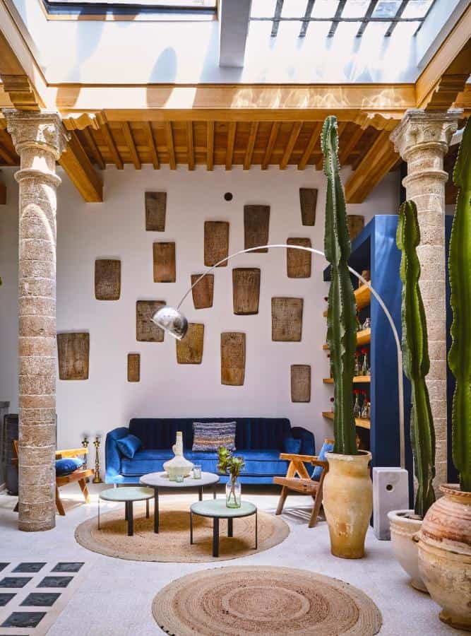 10 BEST Riads in Essaouira 22 A sunlit living room inspired by riads in Essaouira features a blue velvet sofa, tall cactus in clay pots, round coffee tables, rustic columns, wooden ceiling, and wall art. Skylights and earthy decor create a cozy, eclectic atmosphere.