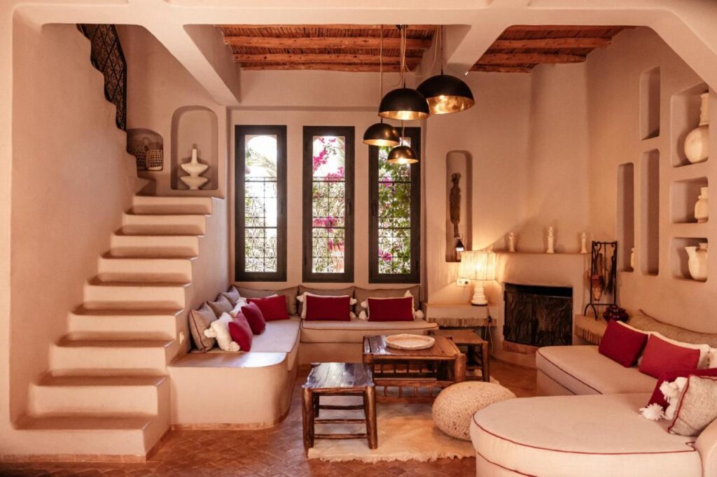 10 BEST Riads in Essaouira 50 A cozy, Moroccan-style living room inspired by riads in Essaouira, with built-in seating, red and white cushions, a wooden ceiling, hanging black lamps, a fireplace, and decorative shelves. Large windows let in natural light and lush greenery outside.