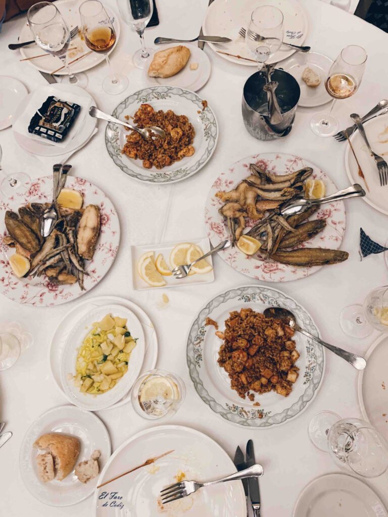 Southern Spain Itinerary 10 Days 15 A table set for a meal with plates of fried fish, rice dishes, lemon wedges, salad, bread, and wine glasses evokes the flavors you'll savor on a Southern Spain itinerary 10 days adventure. Some plates are partially eaten on the white tablecloth.