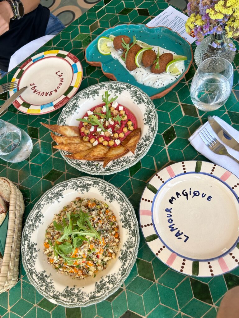 10 BEST Riads in Essaouira 14 A green-tiled table set with colorful Mediterranean dishes—tabbouleh, beet dip with pita chips, falafel, and floral decor—evokes the charm of riads in Essaouira. Two plates read “L’AMOUR SAUVAGE” and “L’AMOUR MAGIQUE.”.