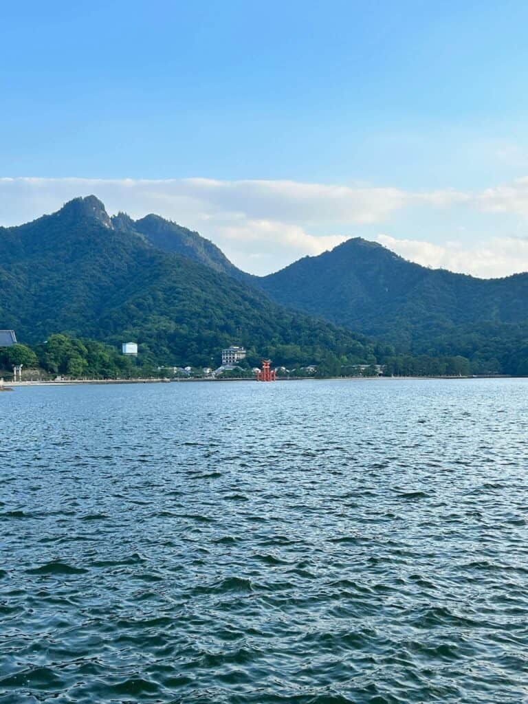 ULTIMATE 10 Day Japan Travel Itinerary 20 A calm body of water with gentle ripples in the foreground, lush green mountains and scattered buildings in the background, and a bright red torii gate near the shore—an iconic scene to include in your 10 day Japan travel itinerary.