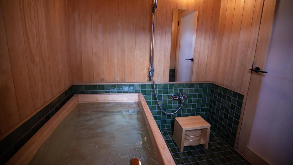 8 UNIQUE Miyajima Ryokans, Hiroshima 38 A wooden Japanese-style bathroom reminiscent of Miyajima ryokans, featuring a deep soaking tub filled with water, green tile walls, a handheld showerhead, wooden stool, and bucket—all reflecting clean, minimalist design.