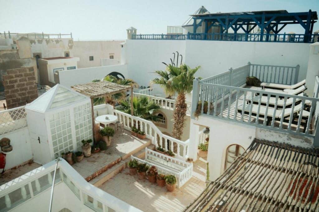 10 BEST Riads in Essaouira 36 A sunny courtyard with white walls, potted plants, palm trees, and shaded pergolas evokes the serene Mediterranean vibe found in riads in Essaouira. Staircases and terraces with railings complete this peaceful retreat.