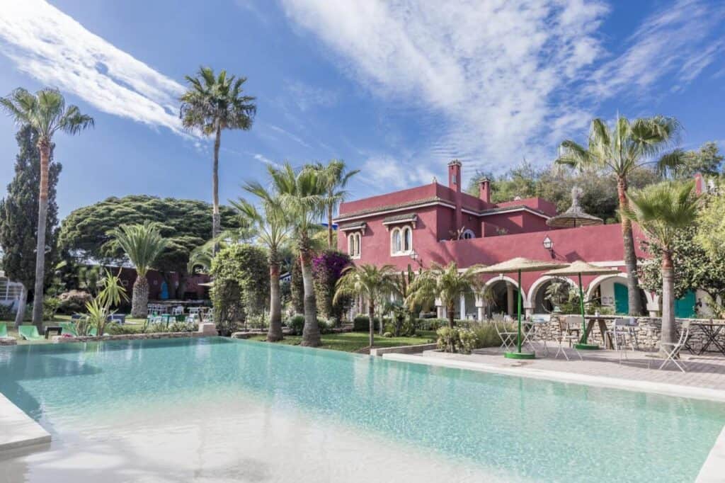 10 BEST Riads in Essaouira 28 A luxurious villa with a red exterior, reminiscent of the elegant riads in Essaouira, is surrounded by palm trees and lush gardens, featuring a large, clear swimming pool under a bright blue sky with wispy clouds.