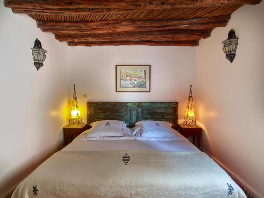 10 BEST Riads in Essaouira 32 A cozy bedroom inspired by riads in Essaouira, featuring a rustic wooden ceiling, a double bed with white linens, two bedside tables with decorative lamps, and framed artwork hanging above the bed.