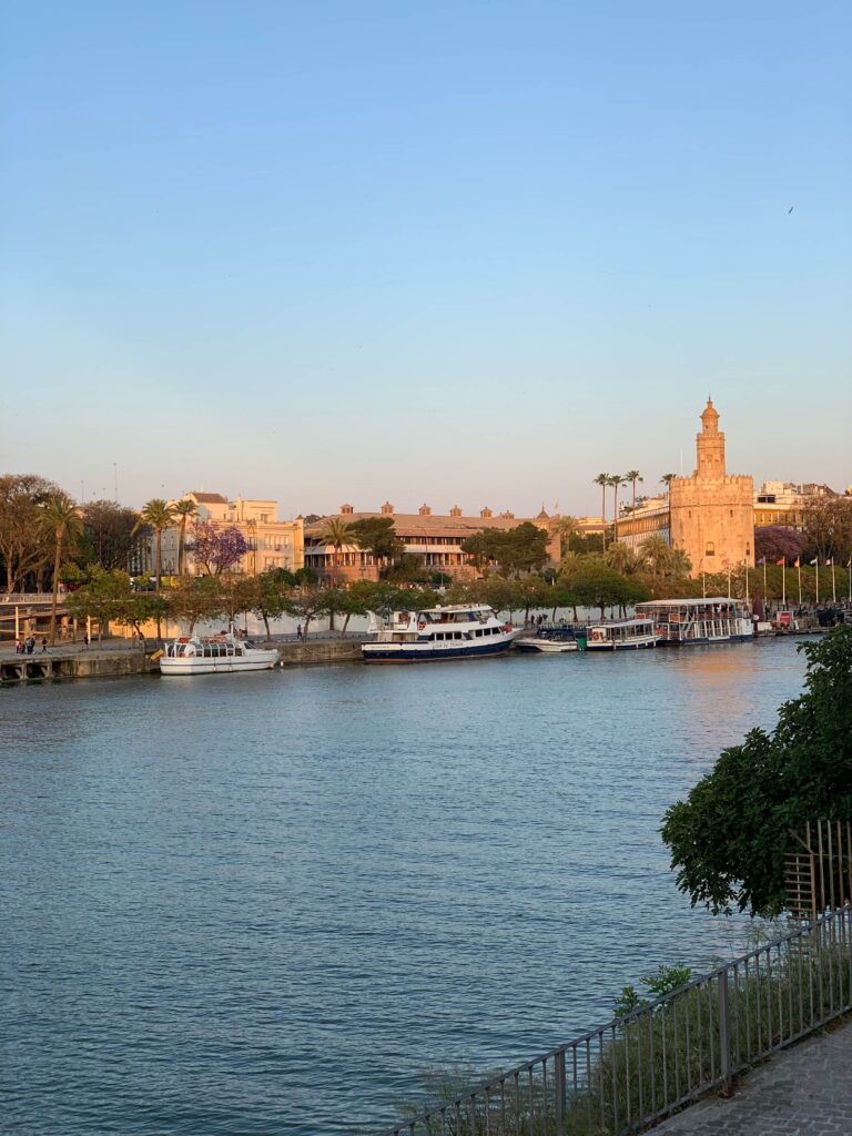 Southern Spain Itinerary 10 Days 13 A calm river with several docked boats, lined by trees and buildings—perfect for your Southern Spain itinerary 10 days. A historic stone tower stands prominently on the right, under a clear blue sky with warm sunlight.