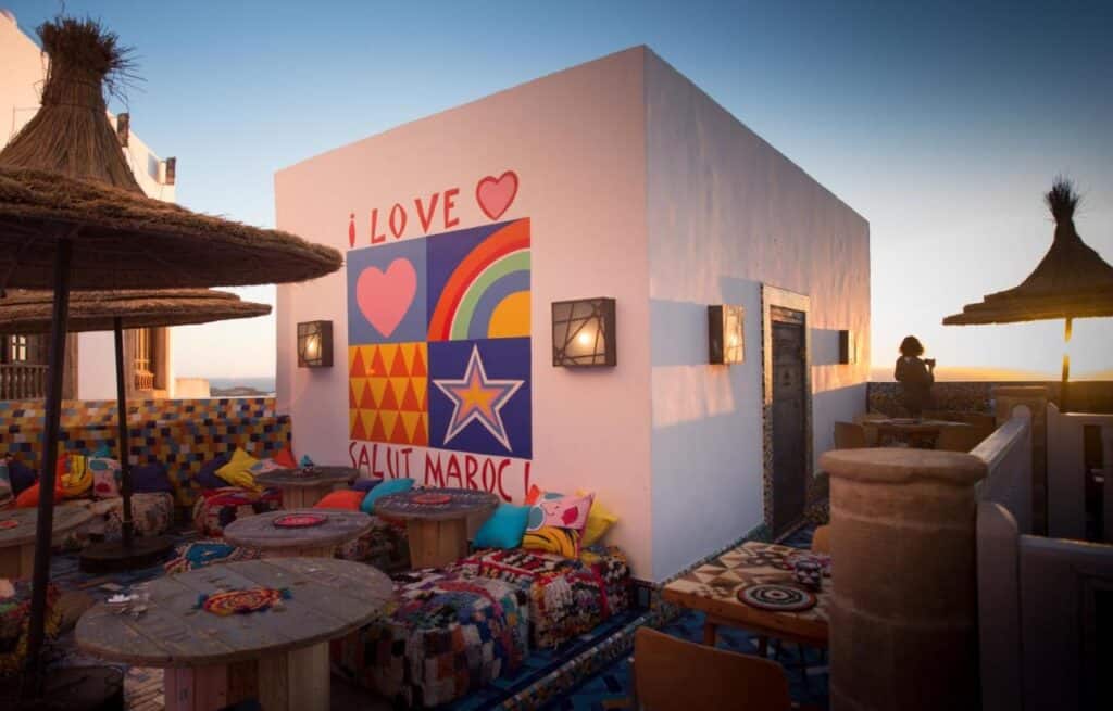 10 BEST Riads in Essaouira 24 Colorful rooftop terrace at one of the riads in Essaouira, with mosaic tables, patterned cushions, and straw umbrellas. A mural reads I LOVE SALUT MAROC in bold shapes. A person stands at the railing, gazing at the sunset.