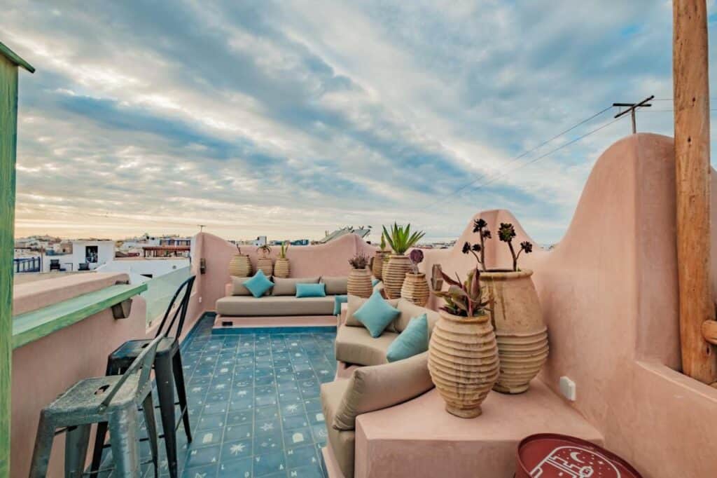 10 BEST Riads in Essaouira 43 A rooftop terrace reminiscent of riads in Essaouira, with blue patterned tiles, beige curved walls, potted plants, cushioned sofas, and metal bar stools overlooks a town under a dramatic cloudy sky at sunset.