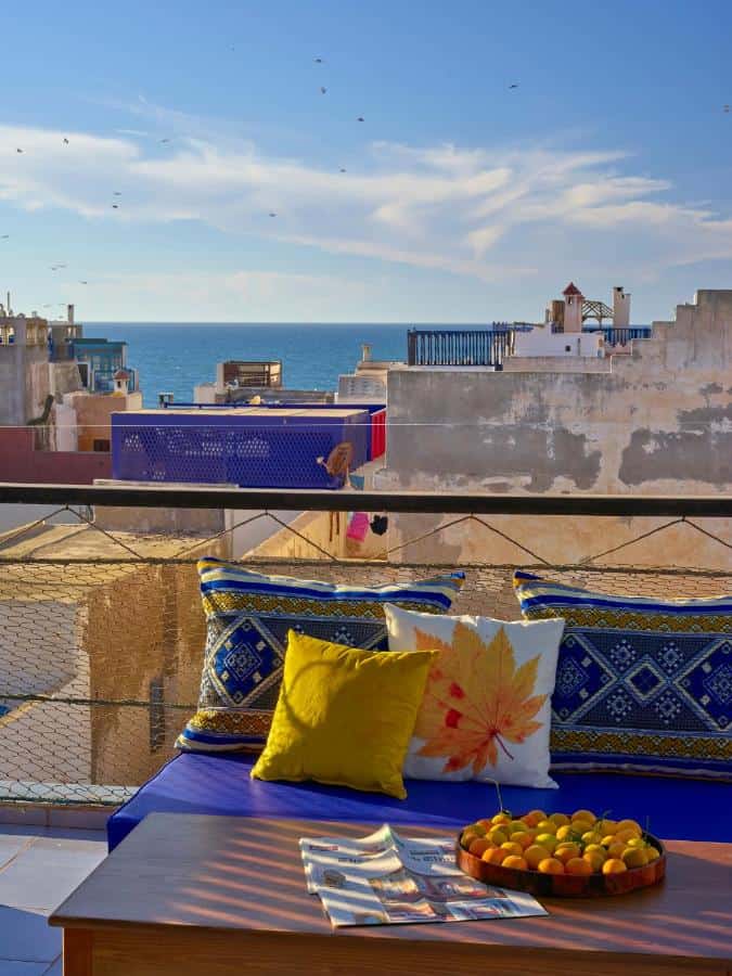 10 BEST Riads in Essaouira 35 A rooftop terrace, reminiscent of riads in Essaouira, with colorful cushions and a table holding fruit and magazines overlooks city rooftops and a distant blue sea under a bright sky.