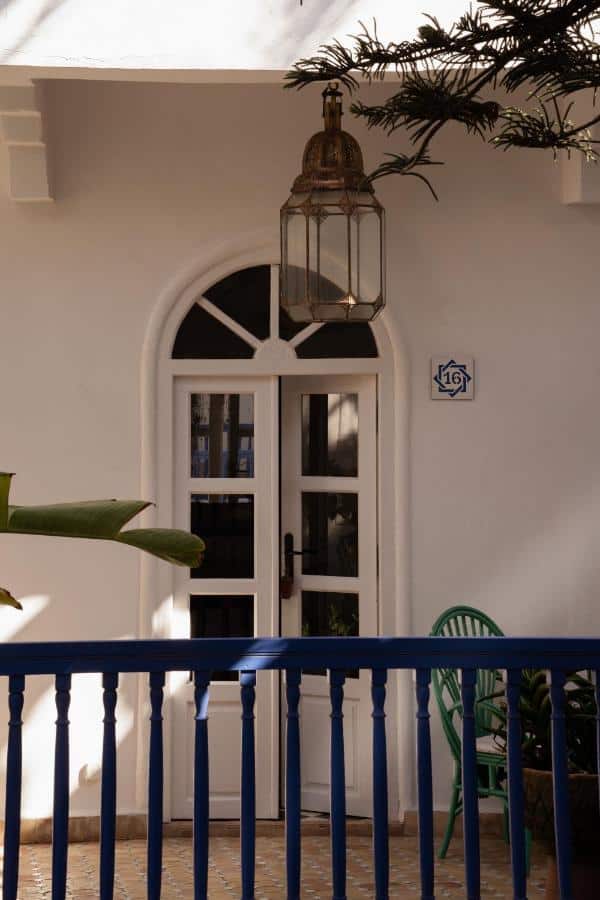 10 BEST Riads in Essaouira 18 A white arched door with glass panes, topped by a large ornate lantern, is framed by a blue railing and green plastic chairs on a tiled patio—capturing the warm, inviting charm often seen in riads in Essaouira.