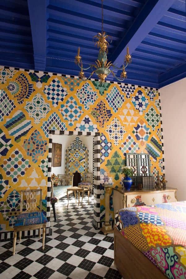 10 BEST Riads in Essaouira 25 A vibrant room, inspired by riads in Essaouira, features bold geometric tile patterns on the walls, a black-and-white checkered floor, bright blue ceiling, ornate gold furniture, and a chandelier. An arched doorway leads to another similarly styled room.