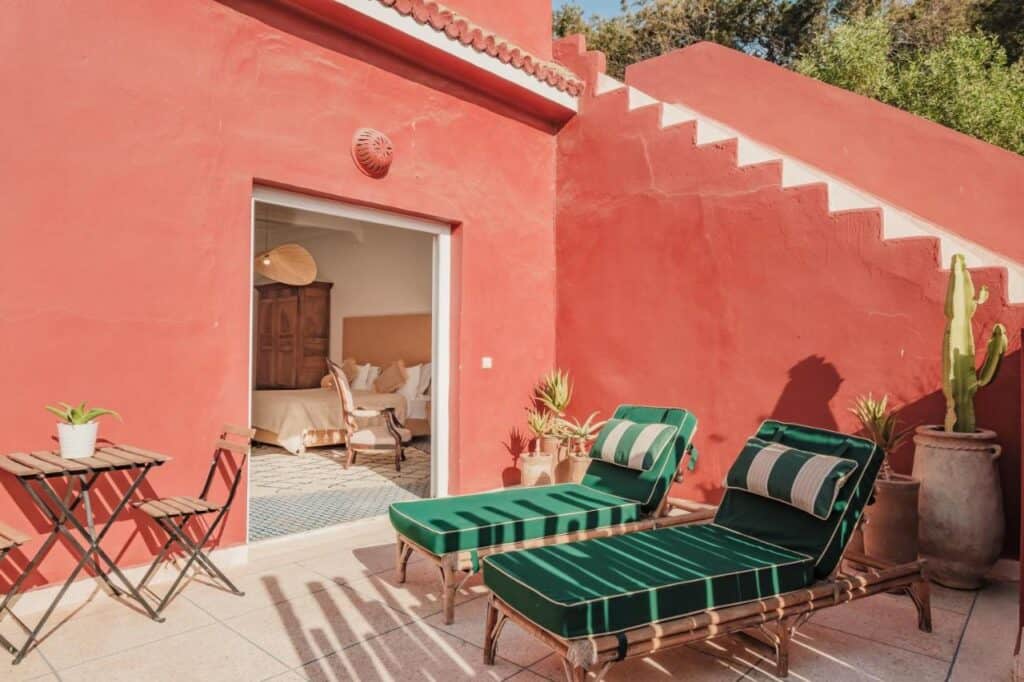 10 BEST Riads in Essaouira 29 A sunlit patio with two green-striped lounge chairs, a bistro table set, and potted plants evokes the charm of riads in Essaouira. Red stucco walls enclose the space, with stairs and a sliding door leading to a bedroom inside.