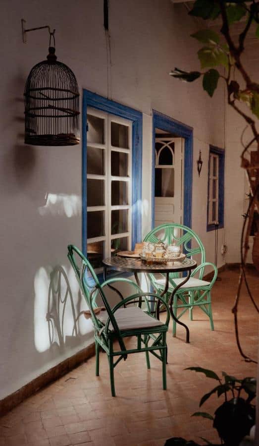 10 BEST Riads in Essaouira 17 A cozy patio, reminiscent of riads in Essaouira, features two green chairs and a round table set for tea against a white wall with blue-trimmed windows. A birdcage hangs above as sunlight casts soft shadows on the tiled floor.